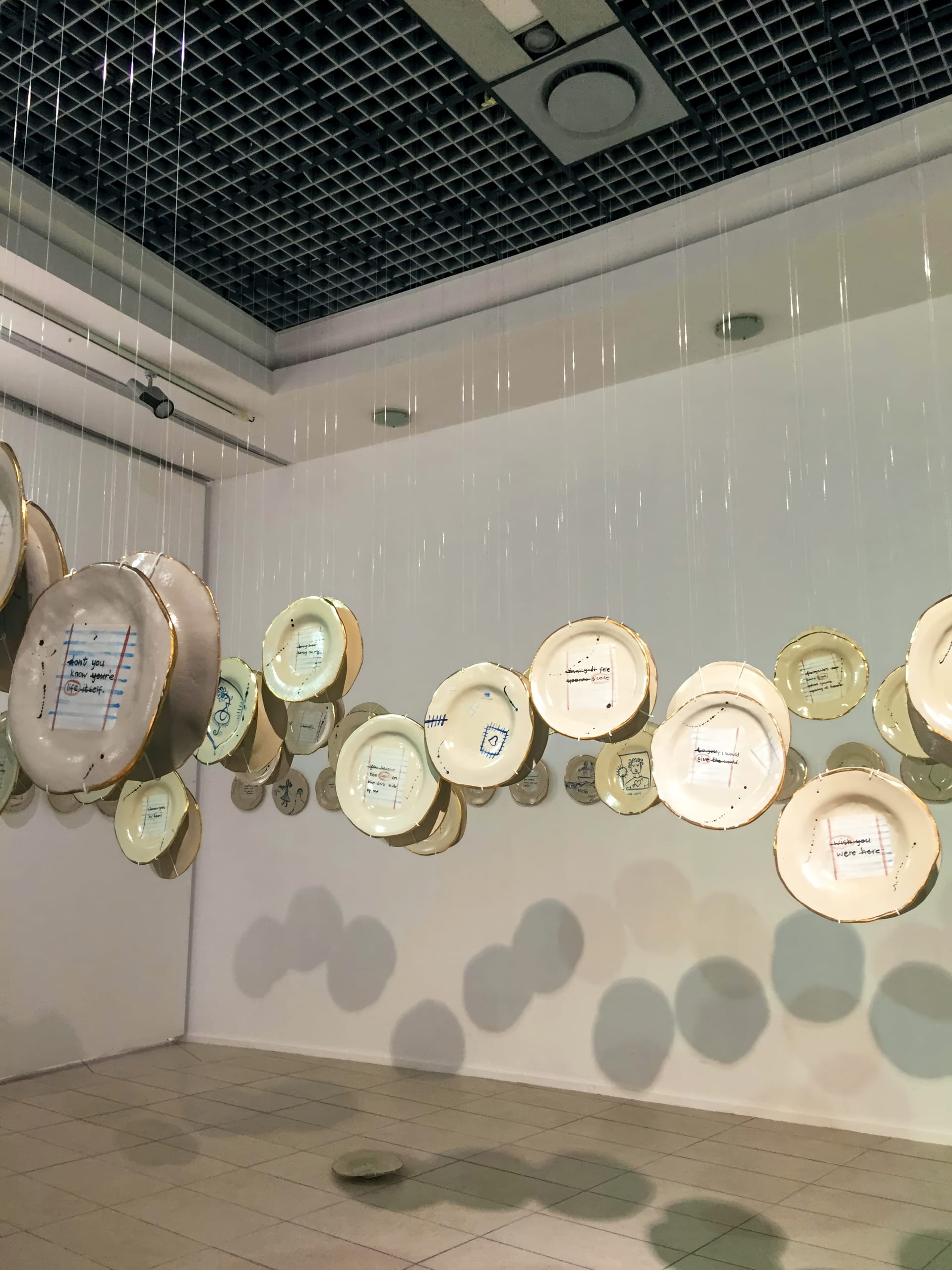 Several ceramic plates with handwritten notes are suspended from the ceiling by clear strings in a gallery space. The plates are arranged at varying heights, casting soft shadows on the tiled floor and white walls. The ceiling has a grid pattern with vents.