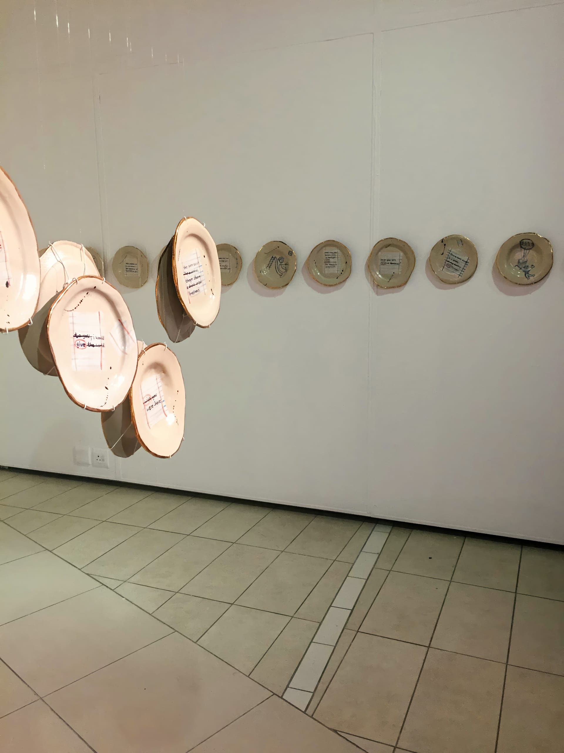 Several embroidered hoops with fabric are displayed on a white gallery wall in a horizontal row, whilst additional hoops hang suspended from the ceiling in the foreground, creating a layered art installation. The setting has tiled floors and minimalist decor.