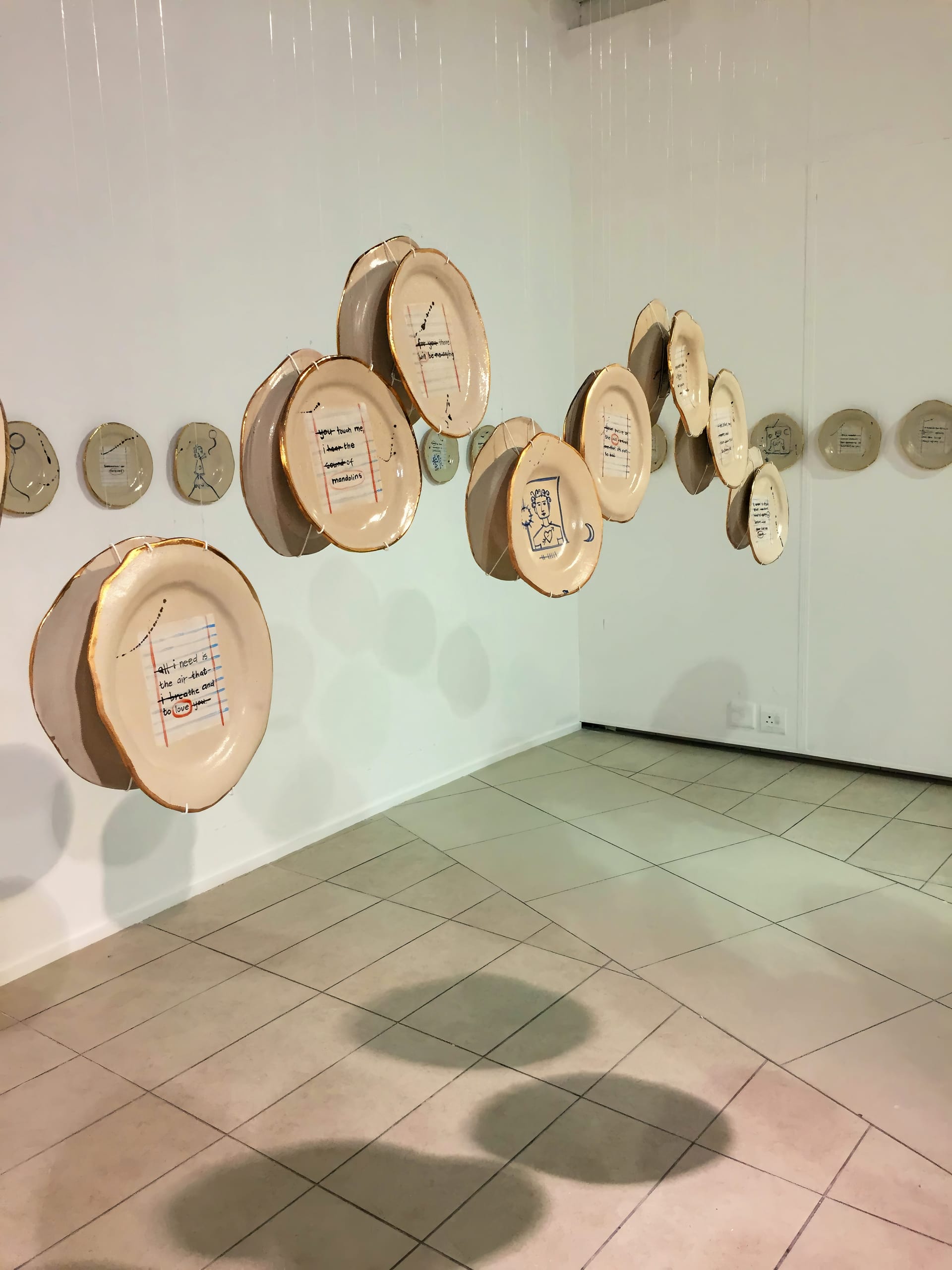Several large, round ceramic plates with artistic drawings and writing are suspended in mid-air in a white gallery space. More plates with similar designs are mounted in a horizontal line along the wall in the background. The floor is tiled.