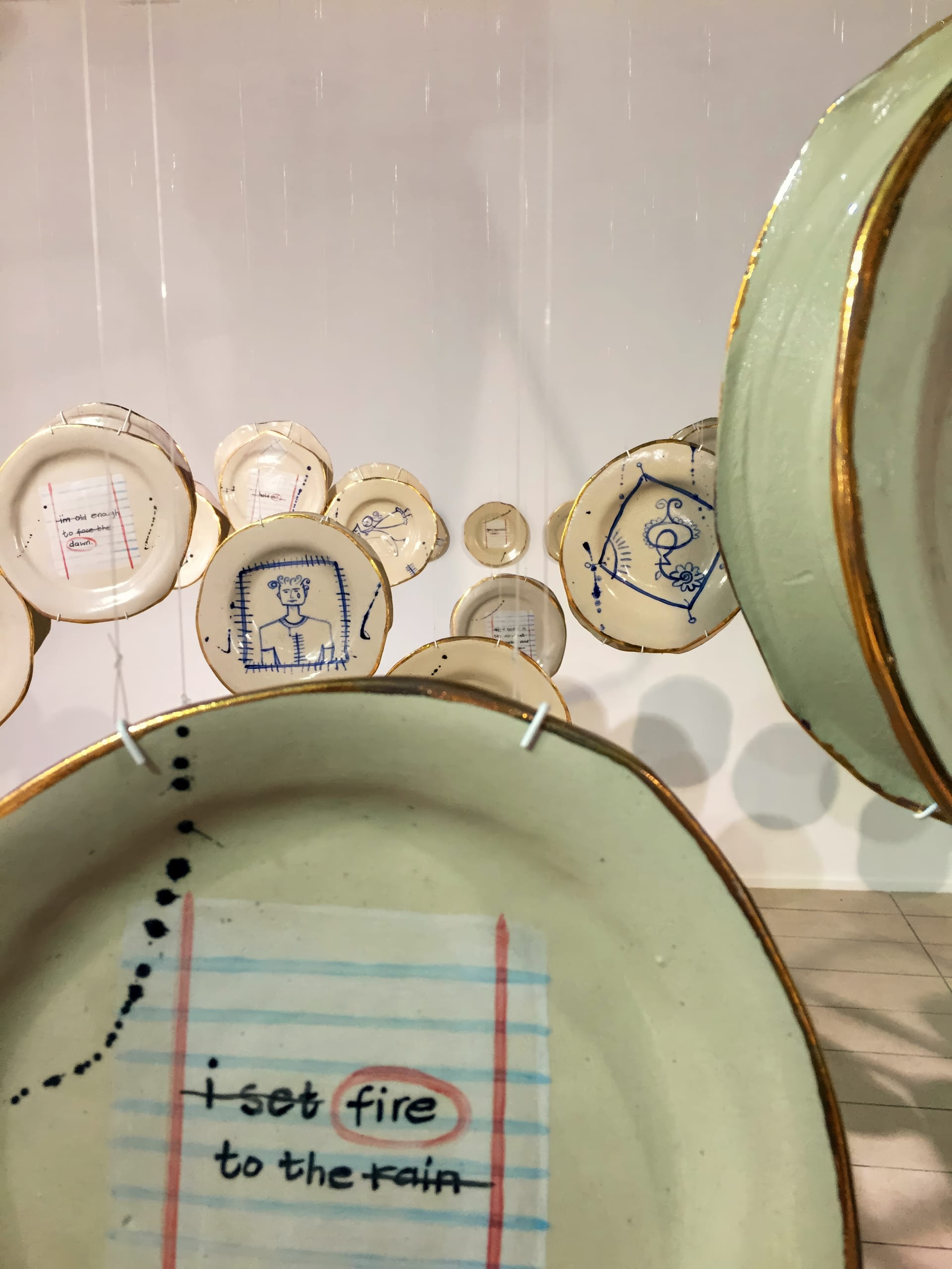 Several cream-coloured ceramic plates with gold rims hang suspended. The nearest plate features a design mimicking lined notebook paper with the handwritten lyrics “i set fire to the rain” in black and blue ink. Other plates show drawings or writing in blue.