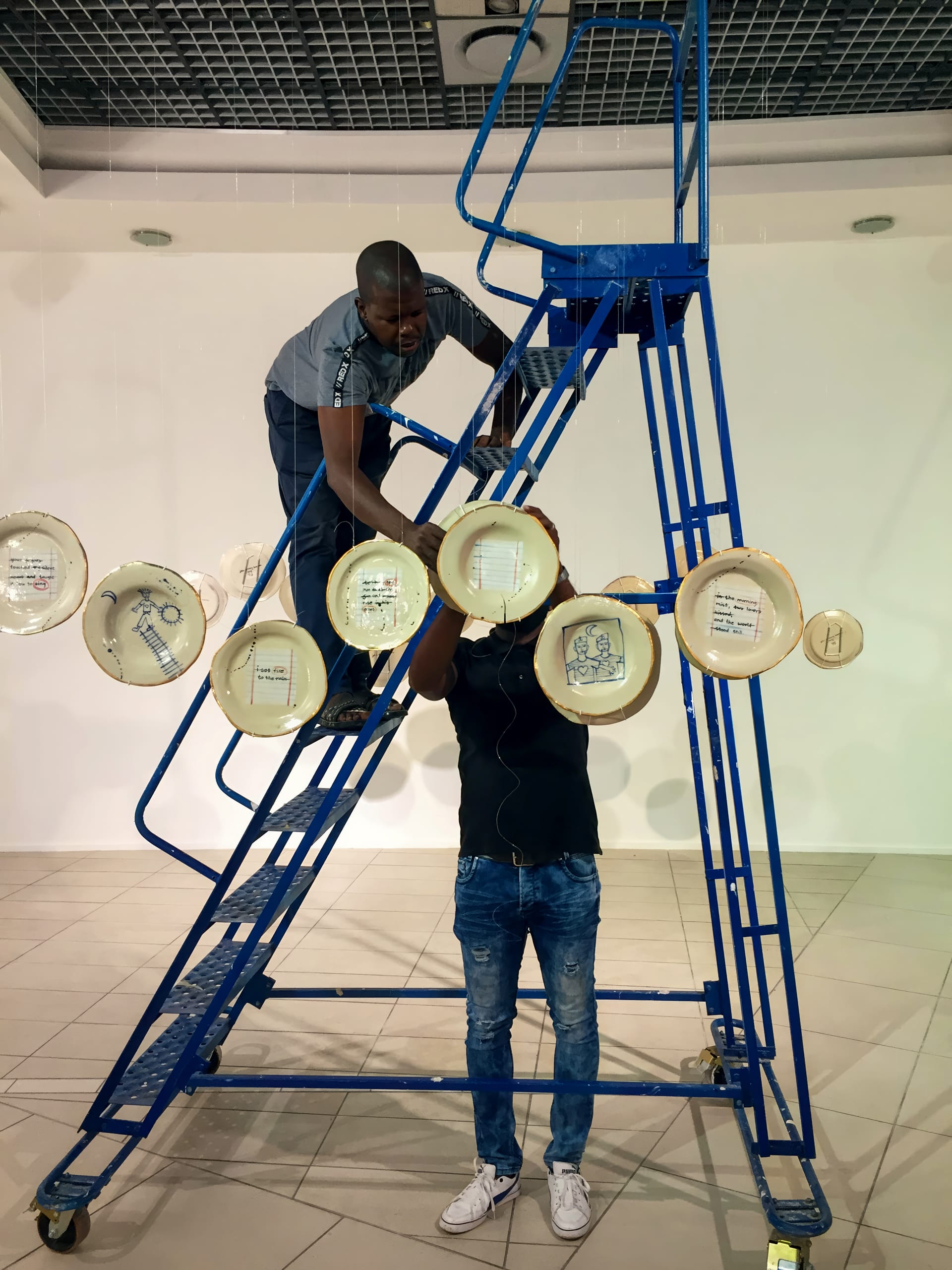 Two people arrange decorative plates on strings hanging from a blue metal ladder inside a bright, tiled room. One person stands on the ladder reaching forward, while the other stands on the floor, partly obscured by the plates.