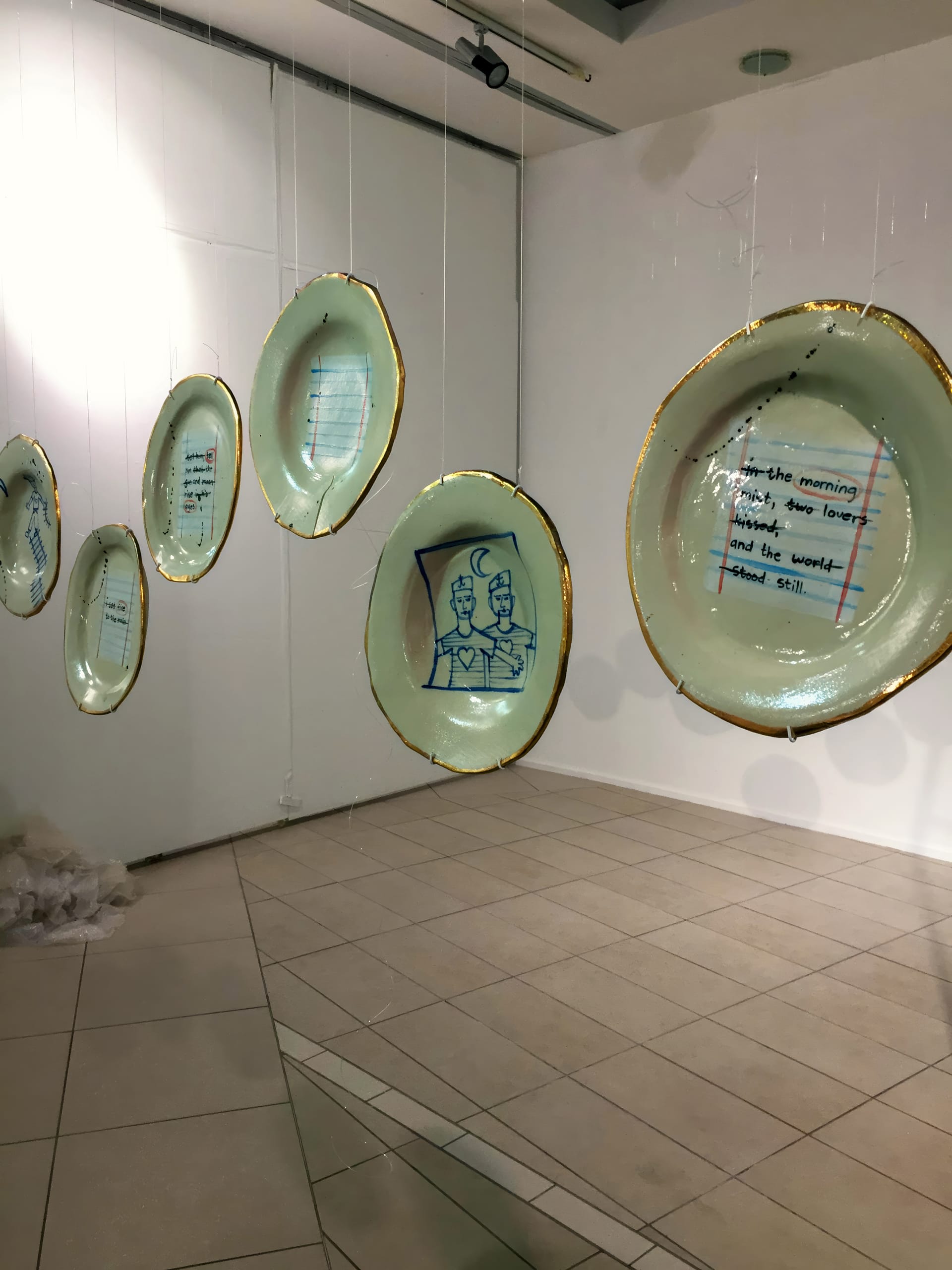 Several large, translucent plates hang in a row from the ceiling in an art gallery. Each plate has gold edges and features blue line drawings or handwritten text, including an illustration of two figures and poetic lines. The plates cast faint shadows on the walls and floor.
