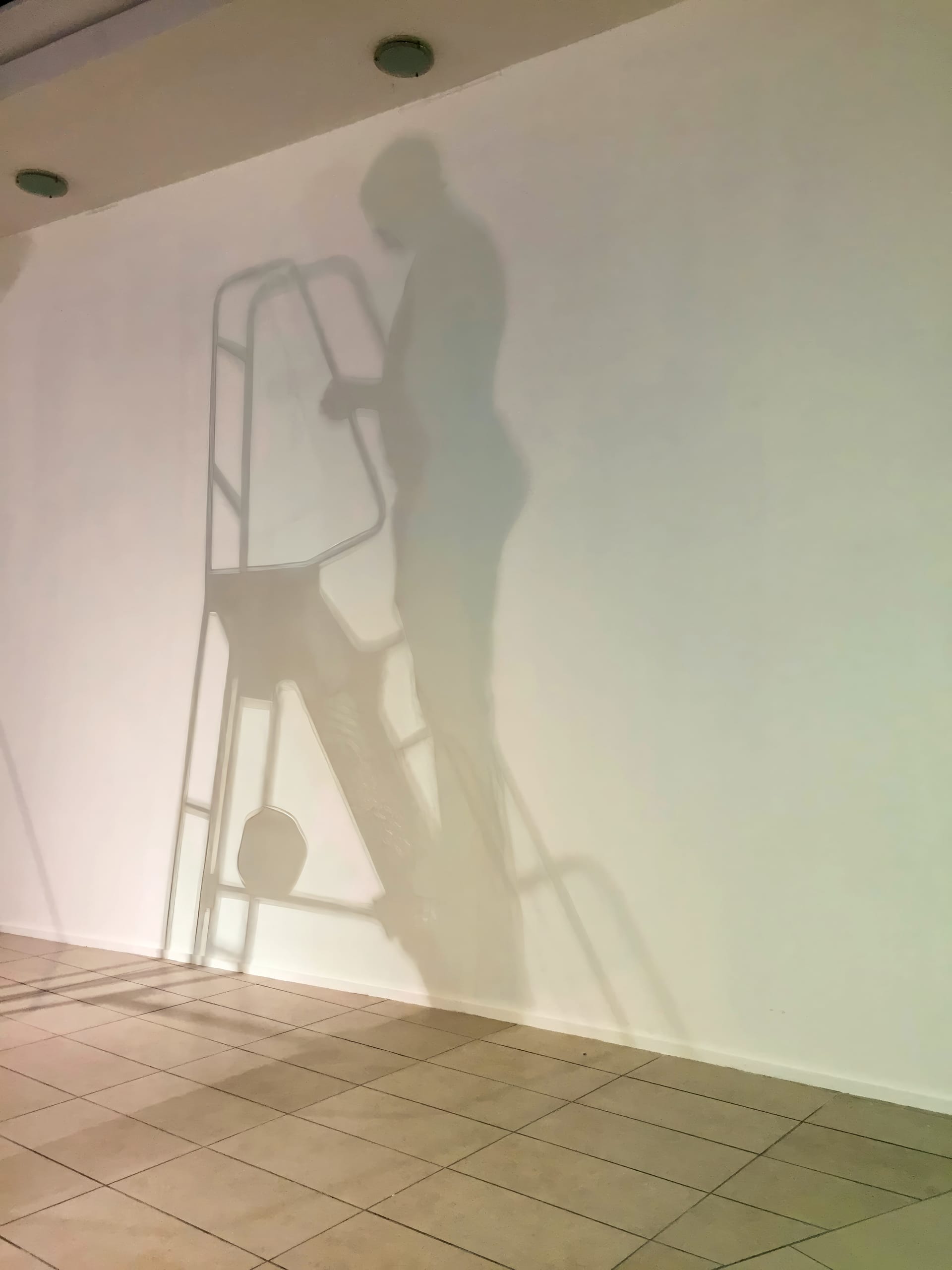 A shadow of a person standing on a step-ladder is cast on a blank white wall. The tiled floor and ceiling lights are also visible, but the actual person and ladder are not shown—only their clear silhouettes.