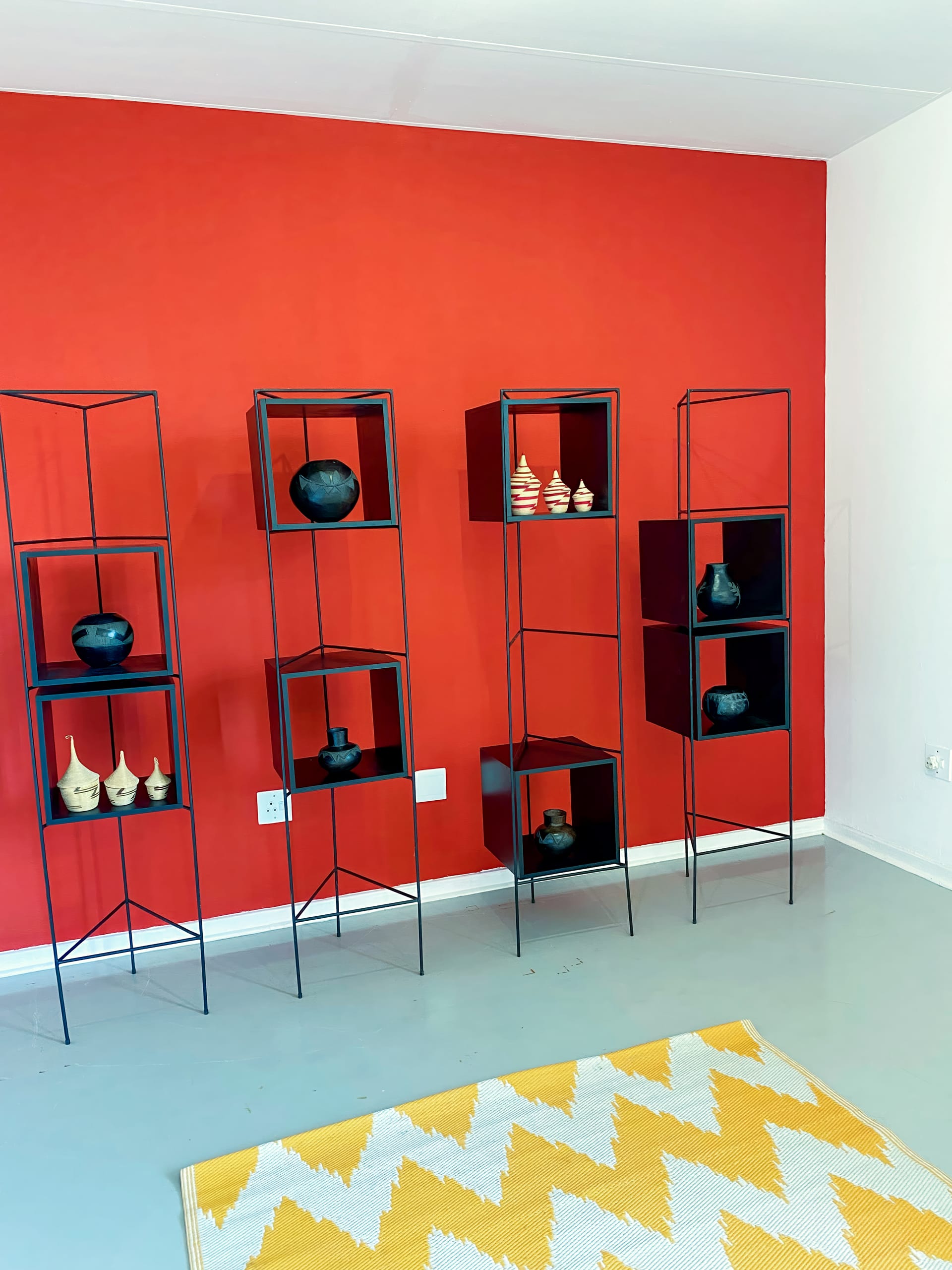 Four black metal display shelves stand against a bold red wall, each holding small black or striped pottery pieces. The floor is grey, and a yellow and white zigzag rug is partially visible in the bottom right corner.