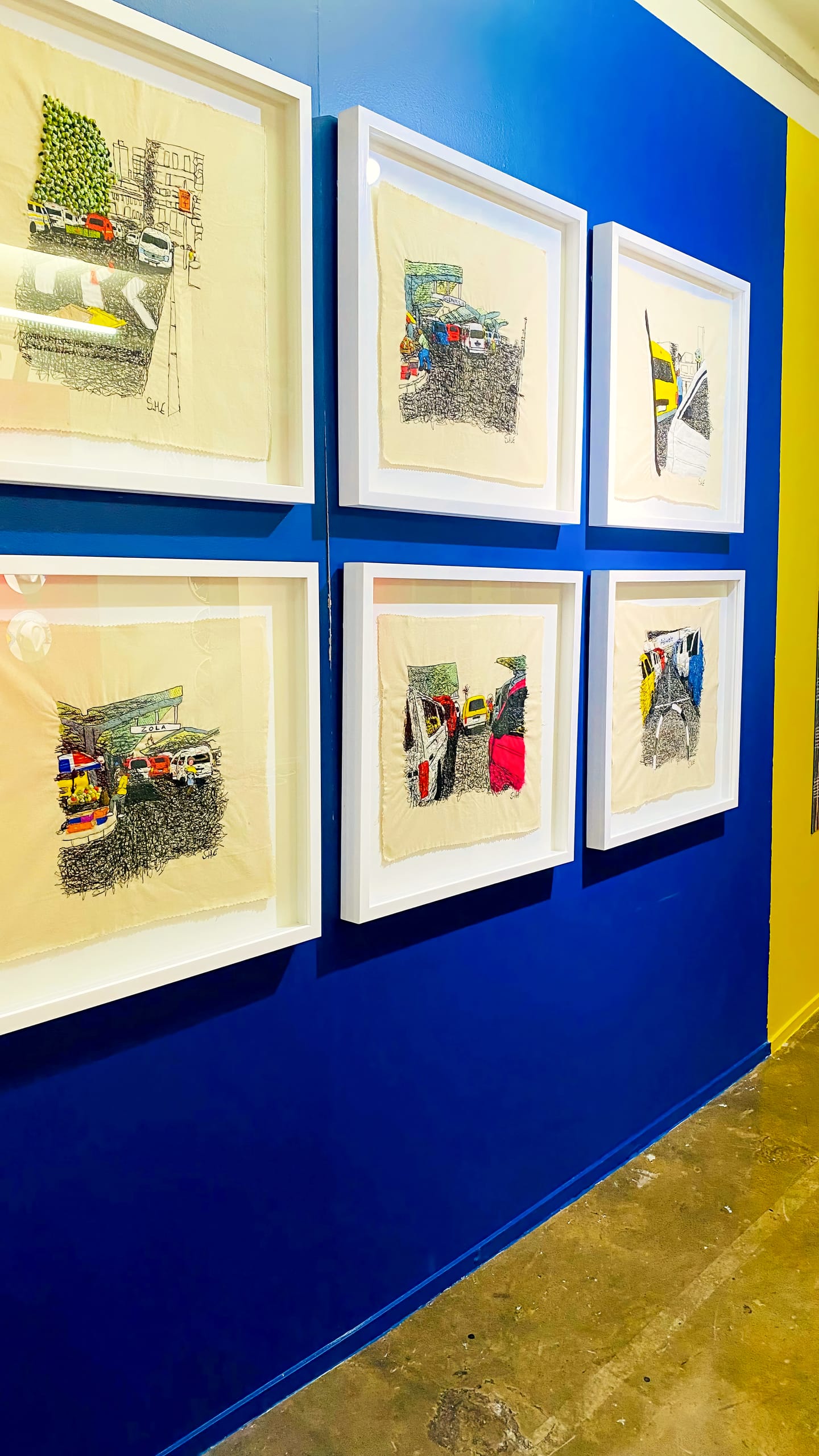 Six framed fabric artworks hang on a blue wall. Each piece depicts a colourful, abstract urban or street scene, possibly featuring cars. The floor beneath is concrete, and part of a yellow wall is visible on the right side.