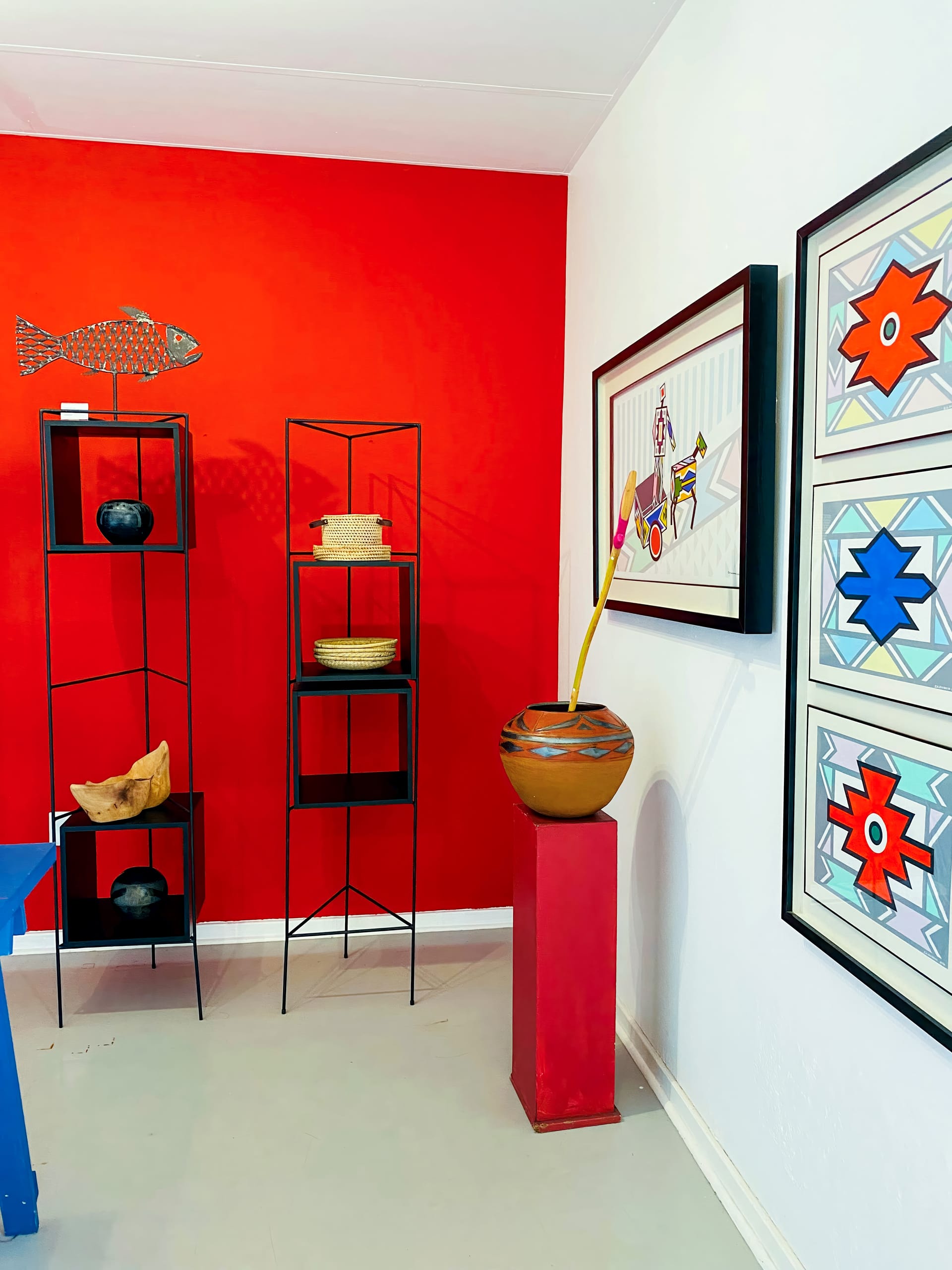 A modern art gallery corner with a bold red feature wall features geometric wire and ceramic art, tall black shelves with pottery, a large patterned vase with decorative sticks, and framed abstract prints with red, blue, and grey designs on a white wall.