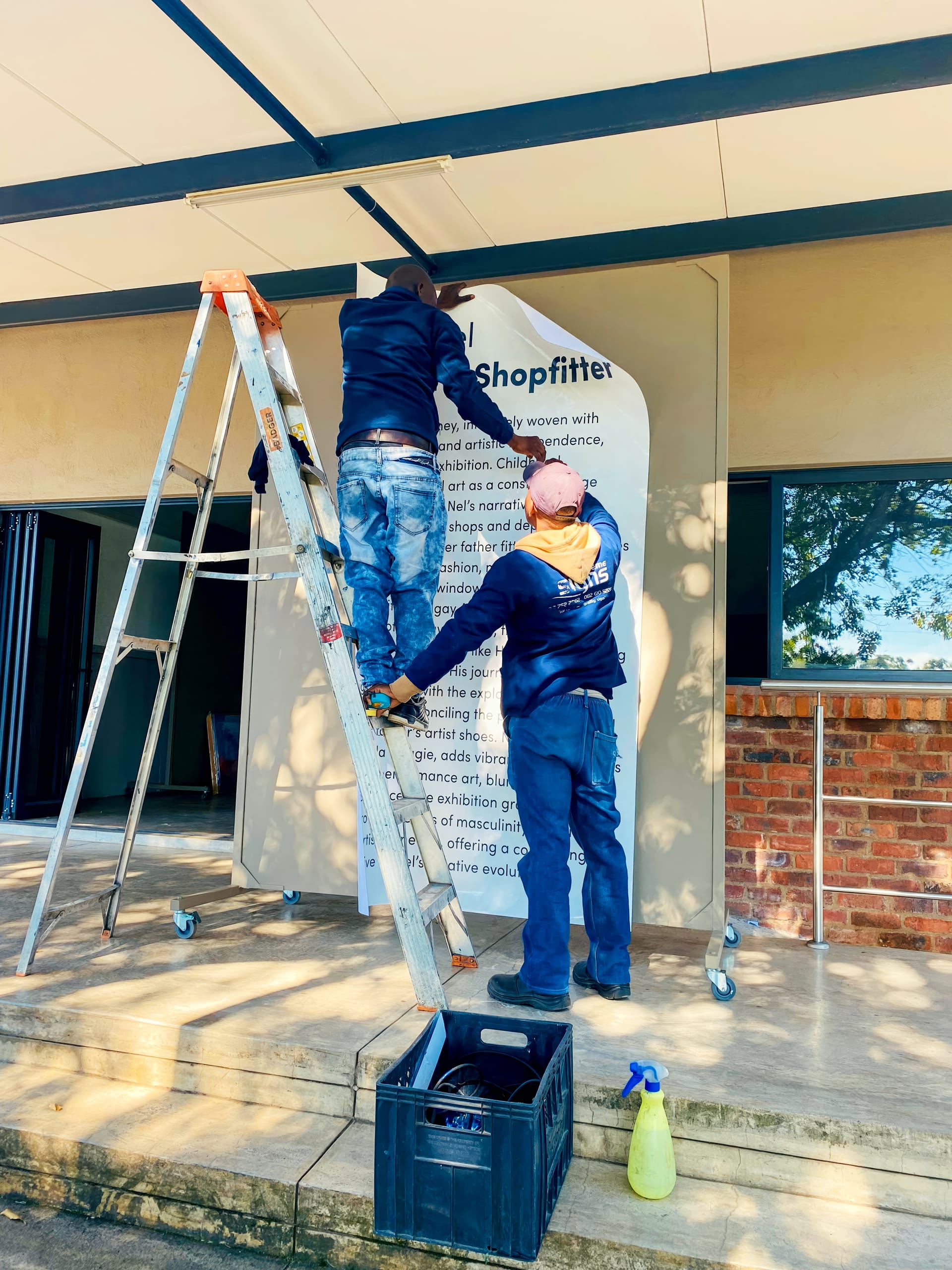 Two men stand on ladders, attaching a large printed poster to a beige outdoor wall. Tools, a crate, and a spray bottle are on the ground nearby. The poster contains text under the heading 
