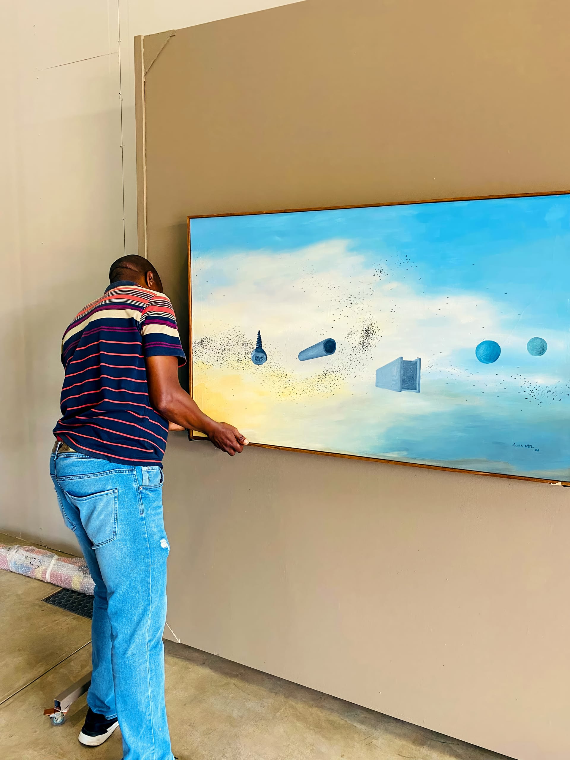 A person in a striped shirt and jeans adjusts a large painting on a beige wall. The artwork features floating geometric shapes such as cones, cylinders, cubes, and spheres against a sky-like, blue-and-yellow background.