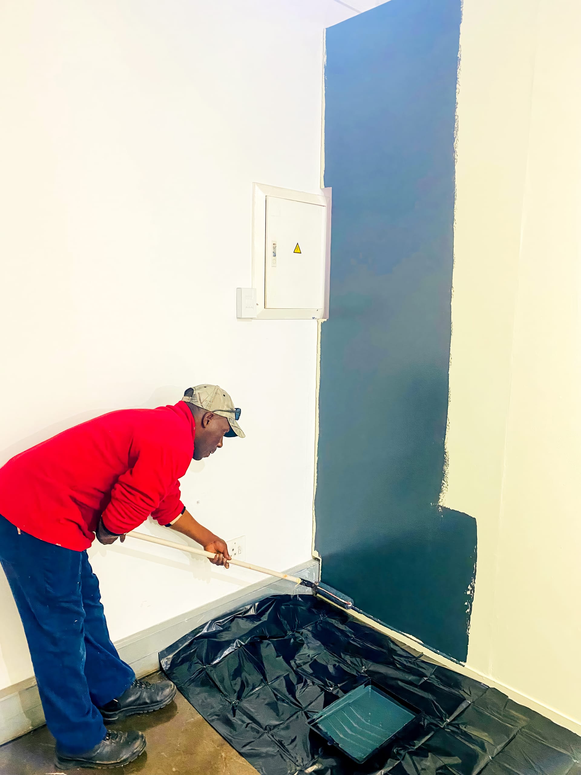 A person in a red shirt and cap uses a roller to paint a white wall dark blue. A plastic sheet covers the floor, with a paint tray on top. The freshly painted section is clearly visible, with uneven edges where the painting has paused.