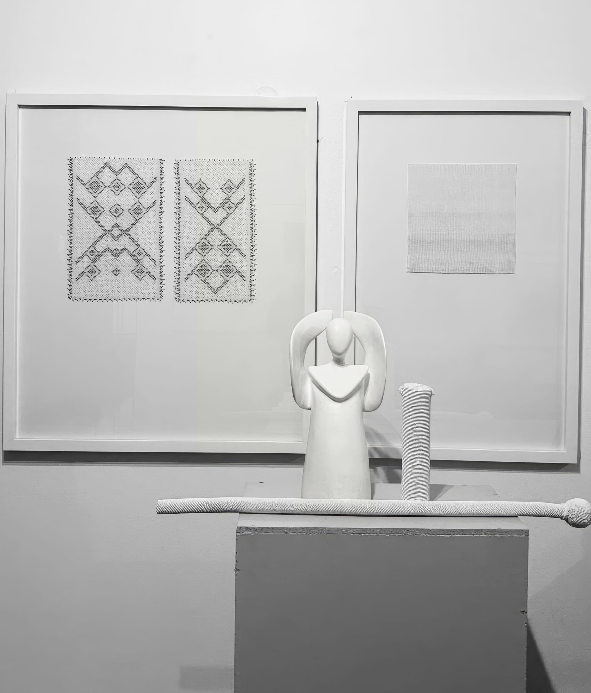 A minimalist art display features two framed geometric textile pieces and a framed gradient square on a wall. In front, a smooth white angel sculpture stands on a plinth beside a textured cylindrical object and a long, rod-like form, all in monochrome tones.