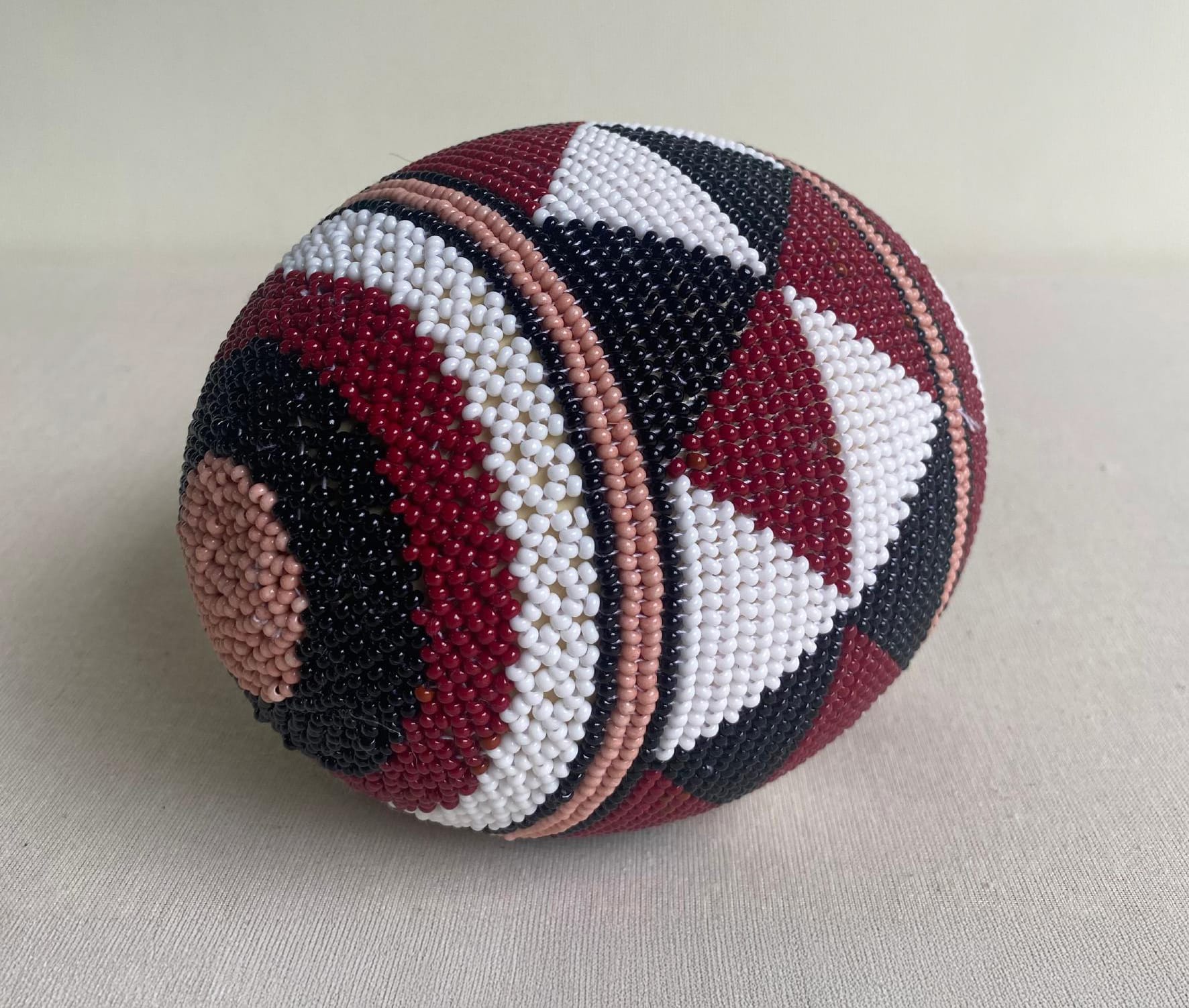 A large, egg-shaped object from Contra.Joburg is covered in intricate beadwork. Geometric patterns in black, white, red, and beige beads form stripes and circles across the egg, giving it a textured appearance that echoes the Weight of Silence.