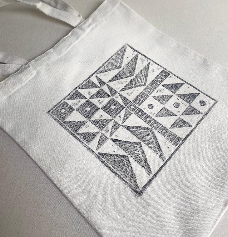 A white fabric tote bag lies flat on a neutral surface. The bag features a large, square geometric design in black ink by Contra.Joburg, displaying patterns of triangles, dots, and lines for an abstract Weight of Silence effect.