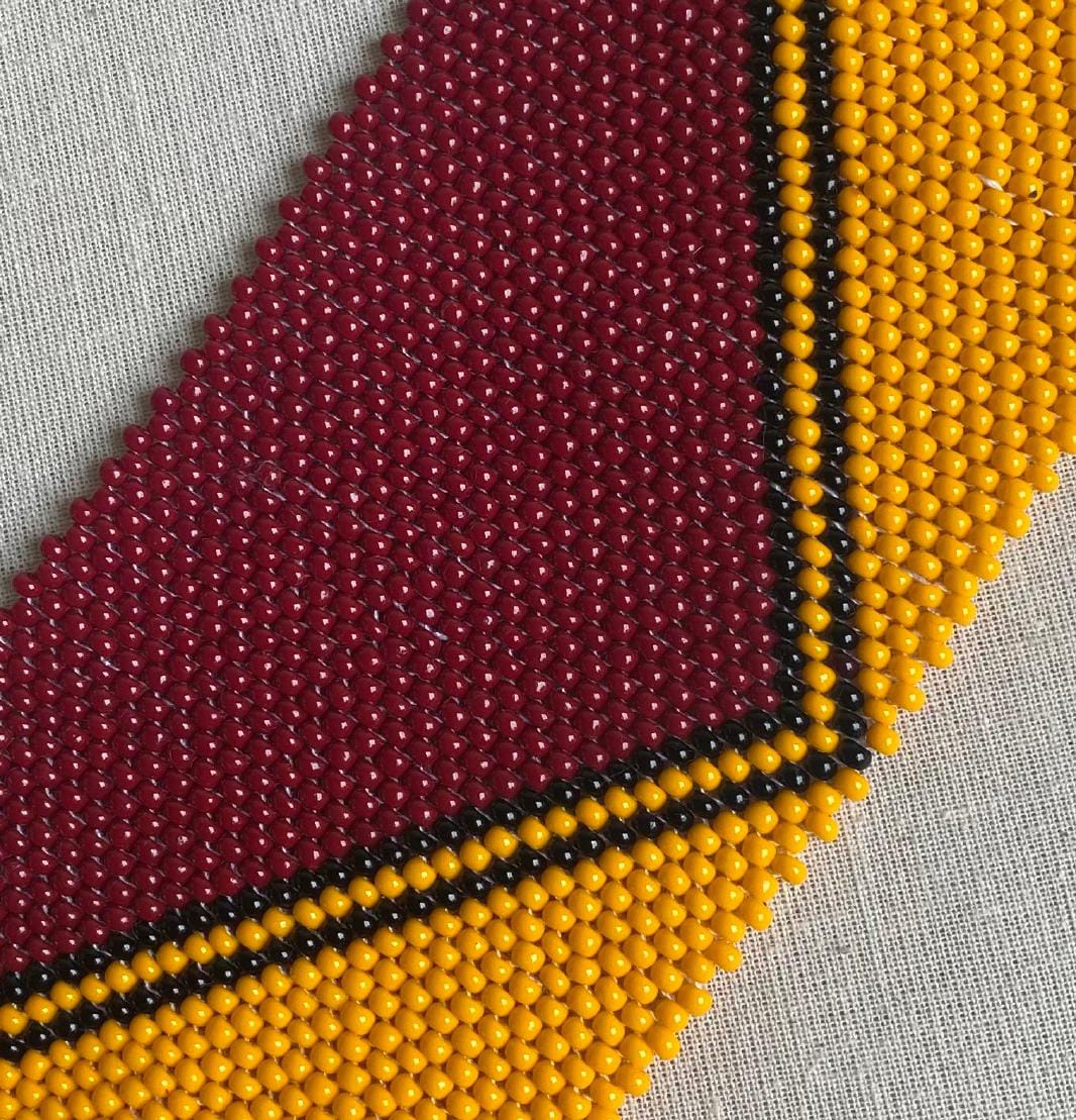 Close-up of a beaded textile from Contra.Joburg with rows of small, glossy beads. The maroon pattern, bordered by black lines and bright yellow beads, evokes The Weight of Silence. Geometric shapes are tightly woven on a light fabric background.