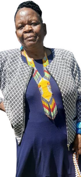 A middle-aged woman stands facing the camera, wearing a navy blue dress and a patterned jacket. She has short hair and a colourful, geometric beaded necklace. Her calm expression reflects the Weight of Silence against a plain, light background.