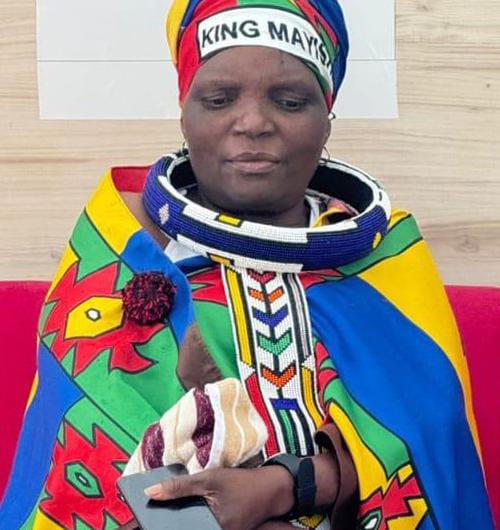 A woman wearing traditional African attire with vibrant, multicoloured patterns and beaded necklaces sits indoors, holding a phone and a small striped cloth. Her headwrap reads 
