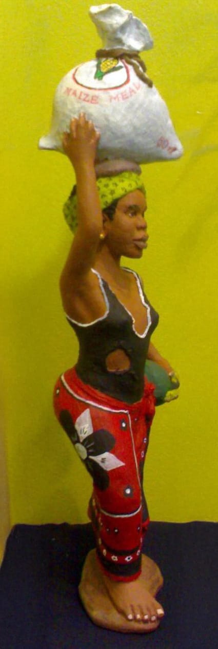 A painted statue of an African woman wearing red patterned trousers, a green headscarf, and a black top with a hole. She balances a sack labelled 