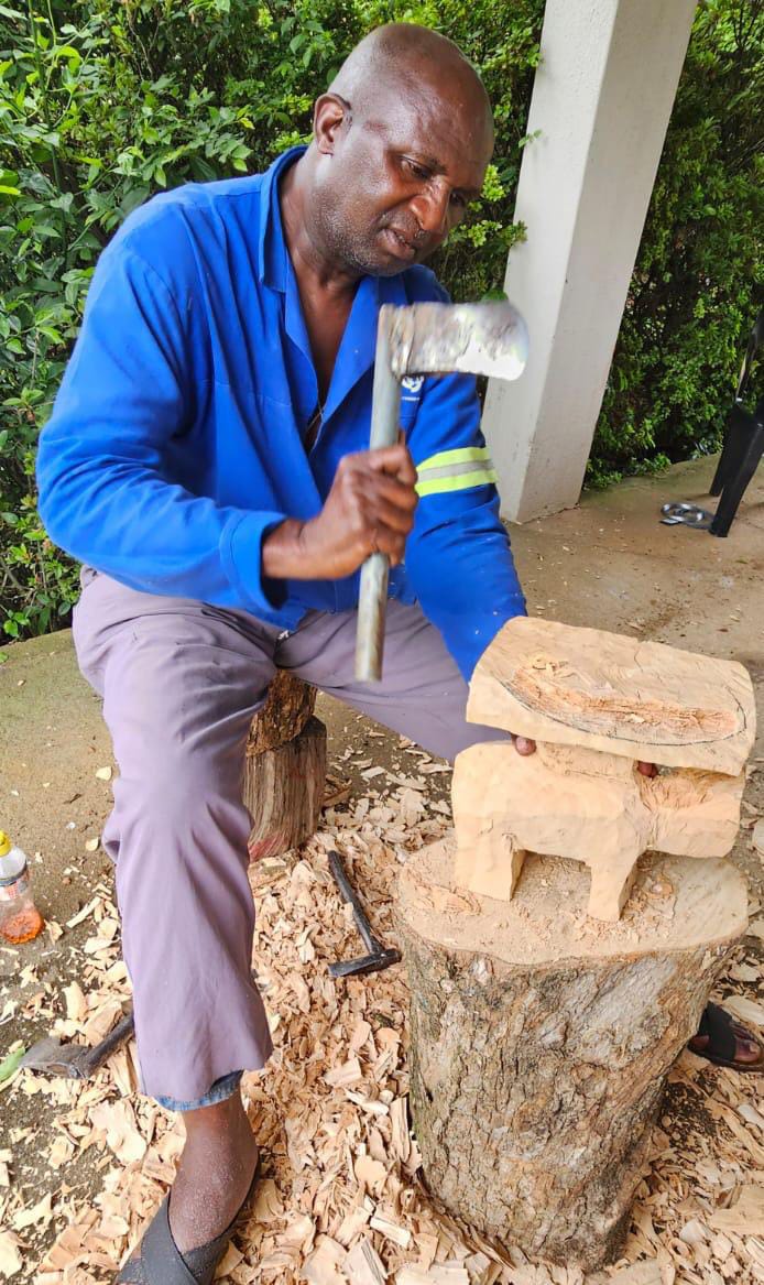 A man in a blue shirt and grey trousers sits on a tree stump outdoors, carving a wooden sculpture with an axe. Wood shavings are scattered around, and green foliage is in the background—a tranquil moment captured by Contra.Joburg.