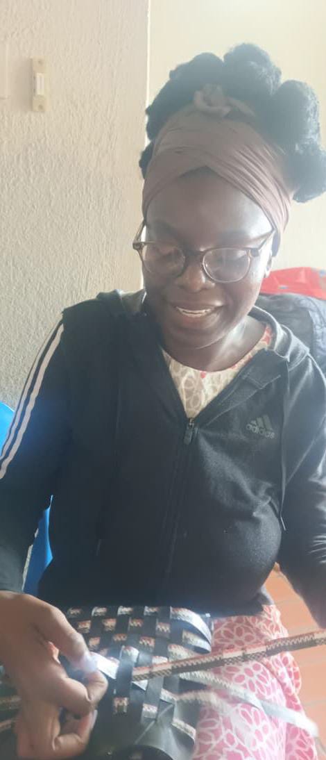 A smiling woman with glasses and a head wrap sits indoors, weaving blue and black plastic strips. Wearing a black Adidas jacket, she is featured in Weight of Silence by Contra.Joburg, focused as soft light illuminates her face.