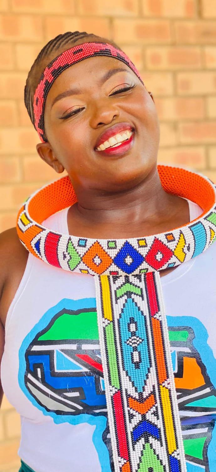 A smiling woman wears a beaded headband and a vibrant, geometric necklace. Her short hair frames a white top displaying an abstract African continent design. Against the brick wall, she embodies the creative spirit of Contra.Joburg.