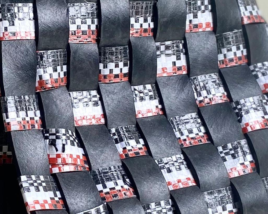 A close-up of an interwoven pattern made of black, glossy strips and metallic tape with a chequerboard design in silver, white, black, and red—echoing the Weight of Silence in its tight, basket-like structure with a slightly curved surface.