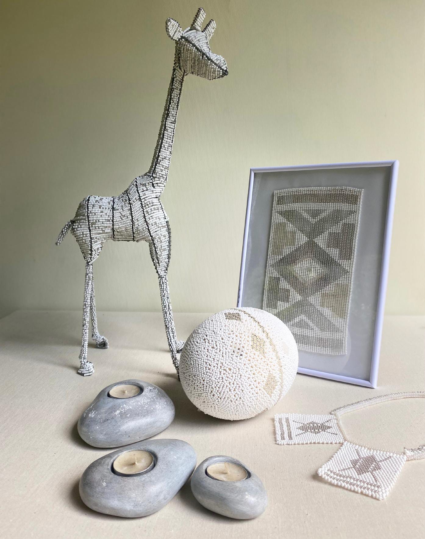 A beaded giraffe sculpture, beaded egg, and framed beadwork from Contra.Joburg sit on a light table. Nearby, smooth grey stone candle holders and a beaded necklace evoke the Weight of Silence in this minimalist, neutral-toned display.
