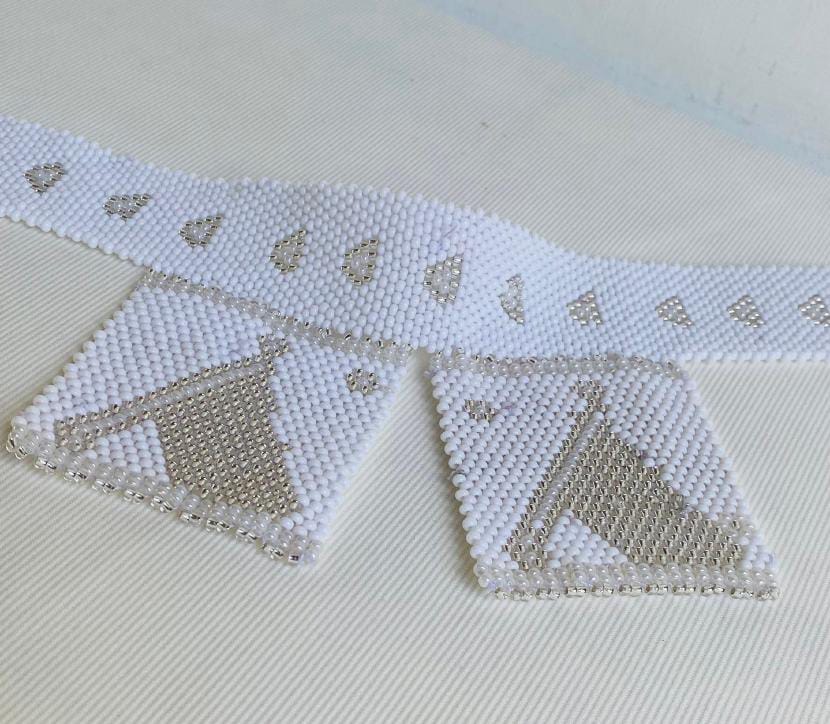 A white beaded belt from the Weight of Silence collection features two pendant-like rectangular sections. Delicate silver and white bead patterns, seen at Contra.Joburg, showcase crescents and abstract designs on a textured white background.