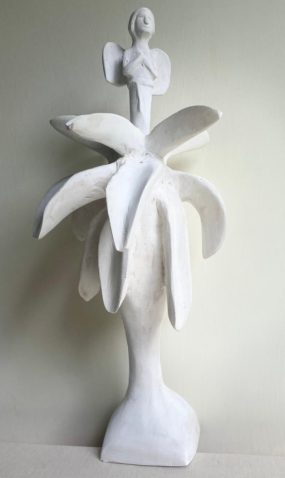 A white abstract sculpture, part of Contra.Joburg’s Weight of Silence series, features a stylised tree with thick branches and a trunk, topped by a winged human figure with outstretched arms against a pale, textured background.