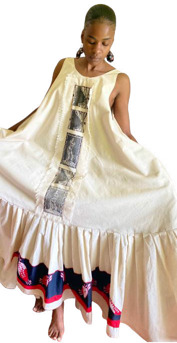 A person wearing a long, sleeveless white dress with a flared bottom and ruffled hem from Contra.Joburg. The dress features vertical decorative panels and a wide navy, red, and white patterned band near the hem. The person is barefoot—a study in the Weight of Silence.