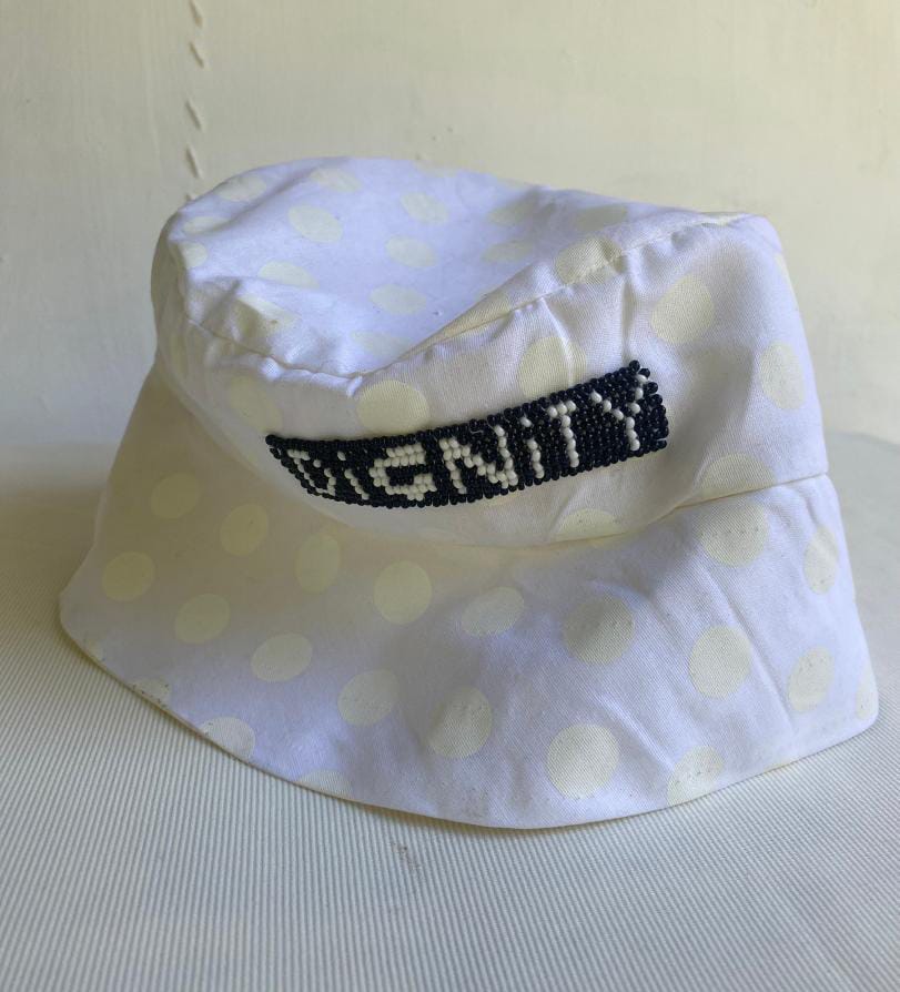 A white bucket hat with large, faint polka dots sits on a white surface. A small rectangular patch with black beaded lettering spells 
