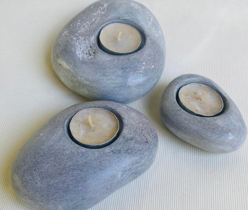 Three smooth, grey stones from Weight of Silence, each with a round cavity holding an unlit tealight candle, are arranged on a light-coloured, textured surface. The stones vary in size and have a natural, matt finish.