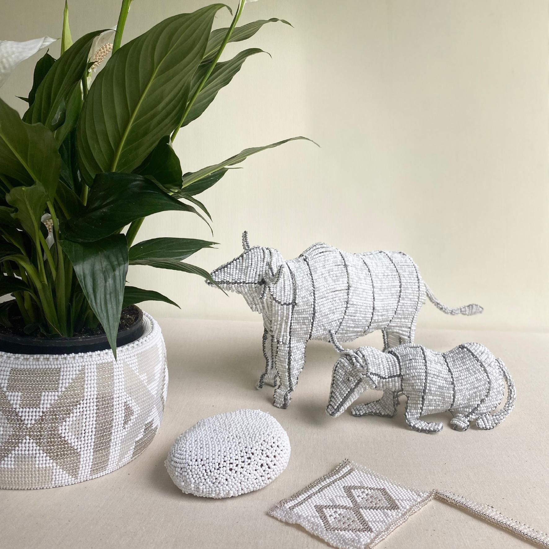 A potted plant with broad green leaves sits in a beaded white and grey pot cover. Beside it, two beaded animal sculptures from Contra.Joburg—a cattle adult and calf—stand near a beaded stone and a pouch patterned like Weight of Silence.