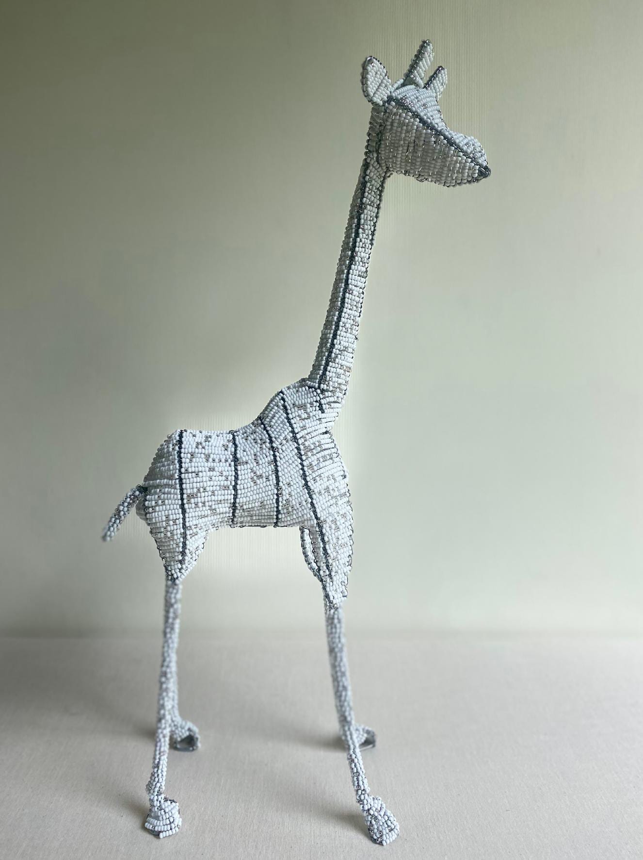 A handmade beaded giraffe sculpture stands on a light surface against a pale background. Crafted with silver and white beads, this Contra.Joburg piece captures the Weight of Silence in its elongated neck, upright ears, and delicately outlined features.
