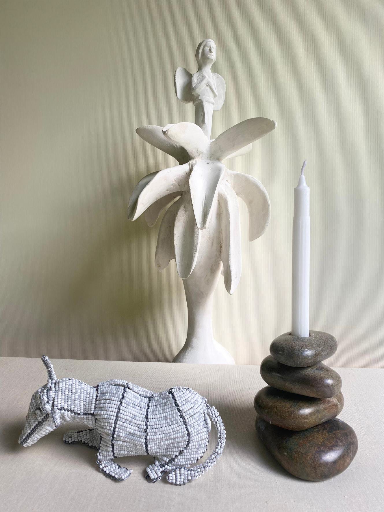 A beaded armadillo figurine rests beside a candle set in stacked smooth stones. Behind them, a white sculpture titled Weight of Silence depicts a woman atop stylised leaves, all set against a light, textured wall.