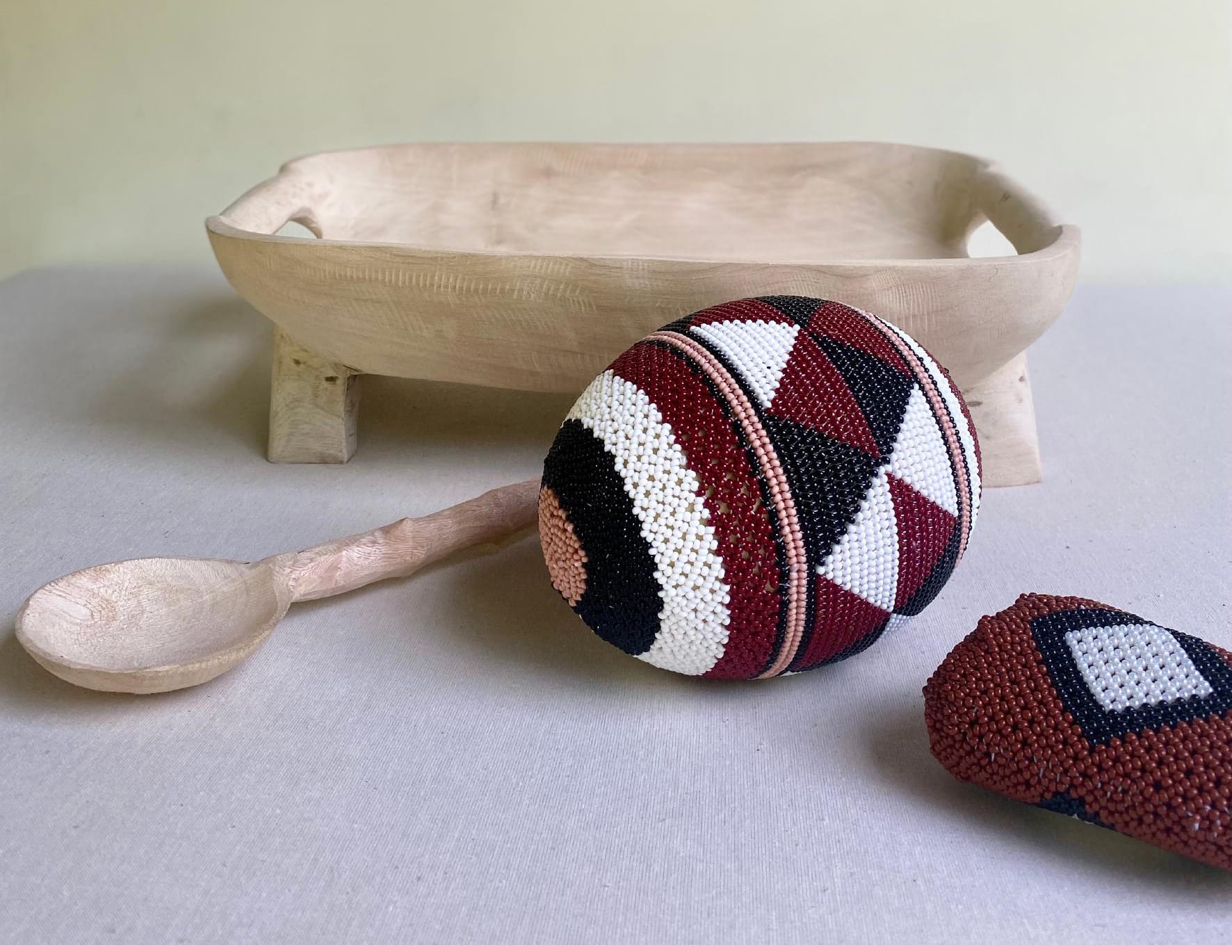 A carved wooden bowl with short legs, a wooden spoon, and two bead-covered ornaments featuring geometric red, black, and white patterns evoke the artistry of Contra.Joburg, displayed on light fabric against a plain, pale background.