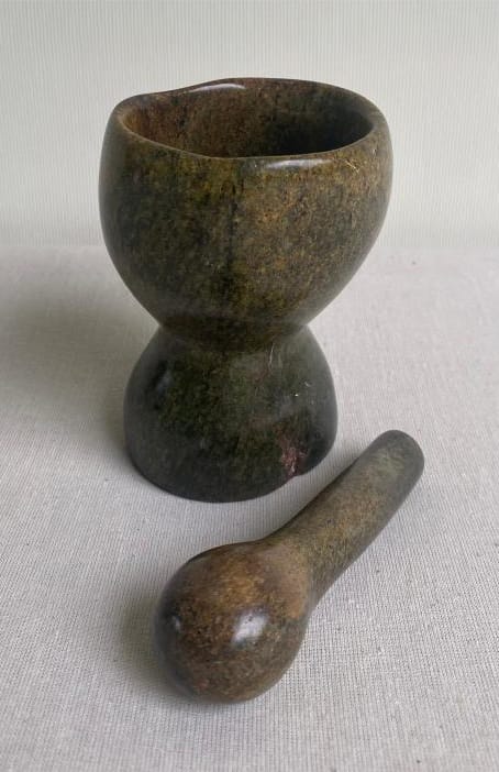 A stone mortar and pestle set sits on a light fabric surface, its smooth, greenish-brown finish evoking the calm Weight of Silence—a fitting accent for any Contra.Joburg-inspired kitchen.