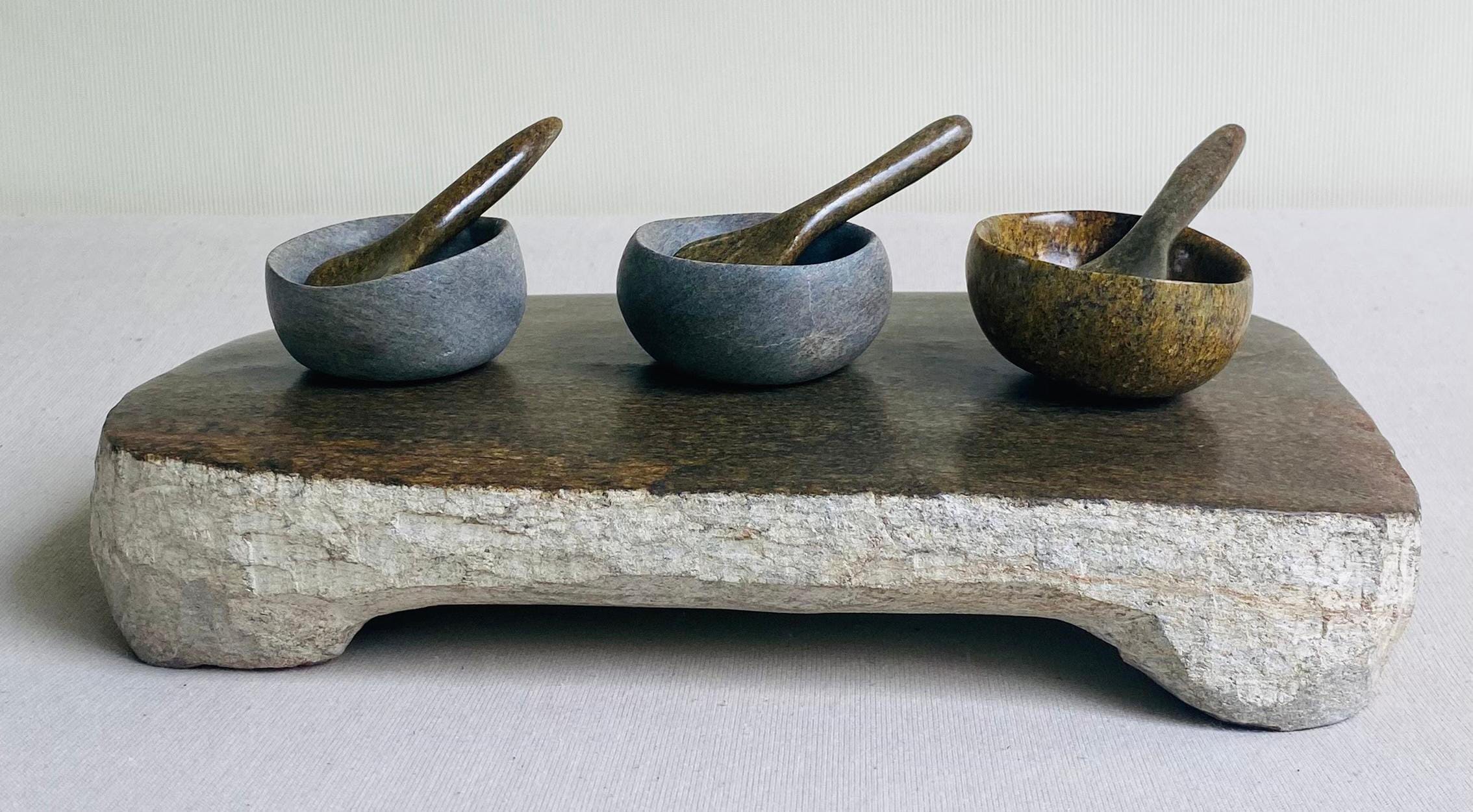 Three small stone bowls with matching spoons sit on a rectangular, rustic slab, evoking the Weight of Silence. Two are grey, one is brown—a minimalist arrangement by Contra.Joburg against a neutral background.