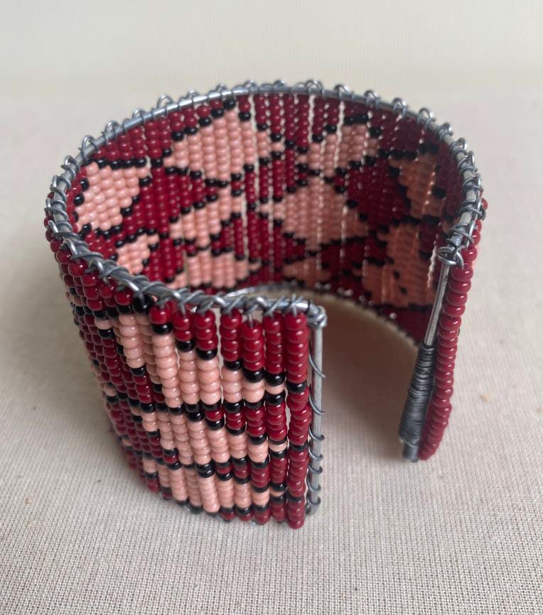 A wide beaded bracelet featuring geometric patterns in red, pink, and black seed beads. Formed around a metal wire frame with visible edges, this Contra.Joburg piece is displayed upright on a neutral fabric background.