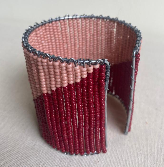 A wide cuff bracelet made of small pink and burgundy beads woven on wire, with the burgundy forming a diagonal stripe. The open back and visible wire edges evoke the artful minimalism found in Contra.Joburg’s Weight of Silence collection.