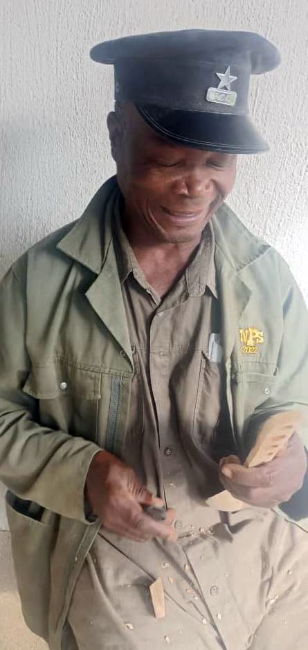 An older man in a uniform with a cap and jacket, smiling whilst whittling wood. Against a light textured wall, wood shavings cover his lap. The star insignia hints at memories—The Weight of Silence quietly present on his embroidered jacket.