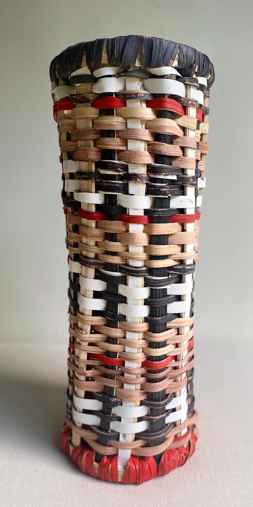 A cylindrical, handwoven basket with a flared rim is featured in this Contra.Joburg piece. Natural, black, red, and white strips form chequered and horizontal patterns. The basket rests on a light surface with a plain background.