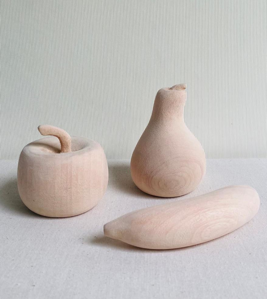 Three wooden sculptures shaped like fruit—a round apple with a curved stalk, a pear standing upright, and a banana lying on its side—sit quietly like the Weight of Silence on a light textured surface with subtle stripes, their smooth pale wood revealing graceful grain.