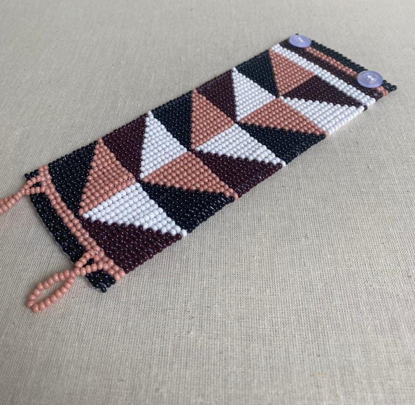 A beaded bracelet with geometric triangular patterns in black, white, mauve, and dark burgundy. The bracelet, featured by Contra.Joburg, has a loop-and-button fastening with two mauve buttons and rests on a textured beige surface.