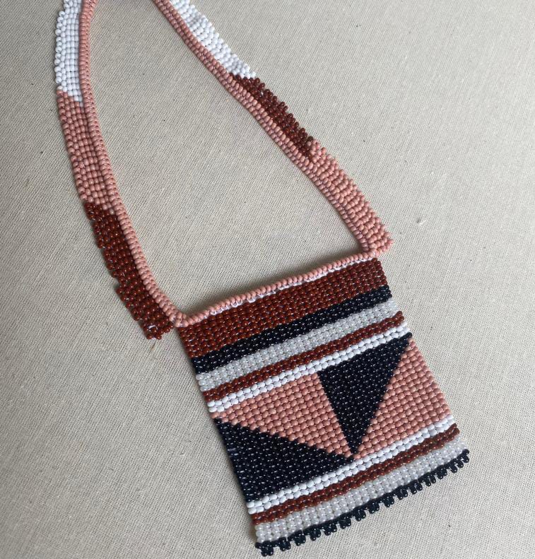 A beaded necklace from Contra.Joburg with a rectangular pouch, featuring geometric patterns in dusty pink, white, black, and dark brown beads. Named Weight of Silence, it’s displayed on a cream fabric background.