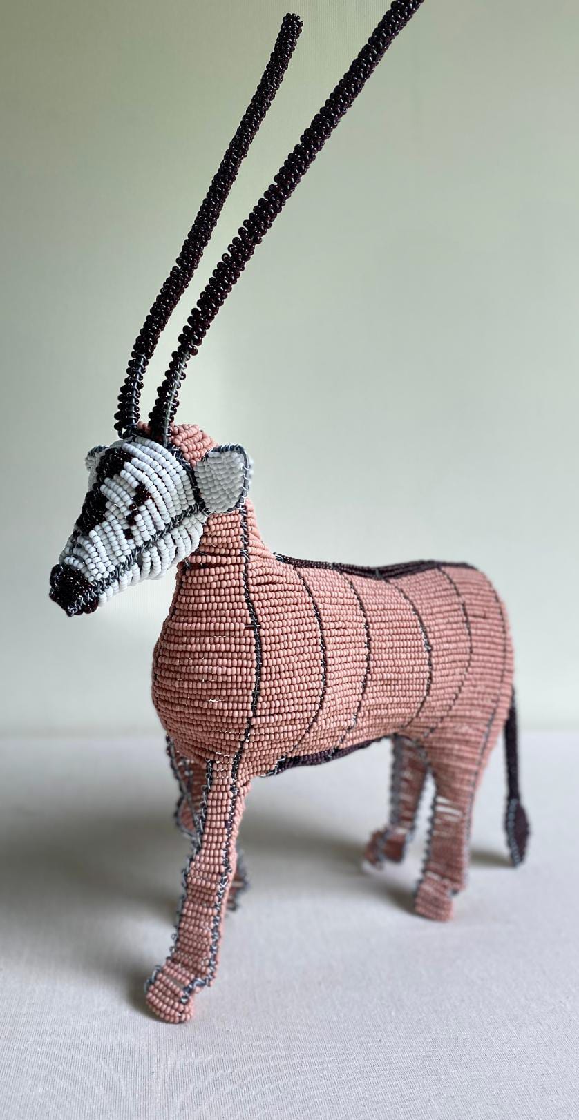 A beaded sculpture of an antelope, featured at Contra.Joburg’s Weight of Silence, stands on a white surface. It has a pinkish body with black stripes, a grey and black face, and two long black beaded horns against a plain background.