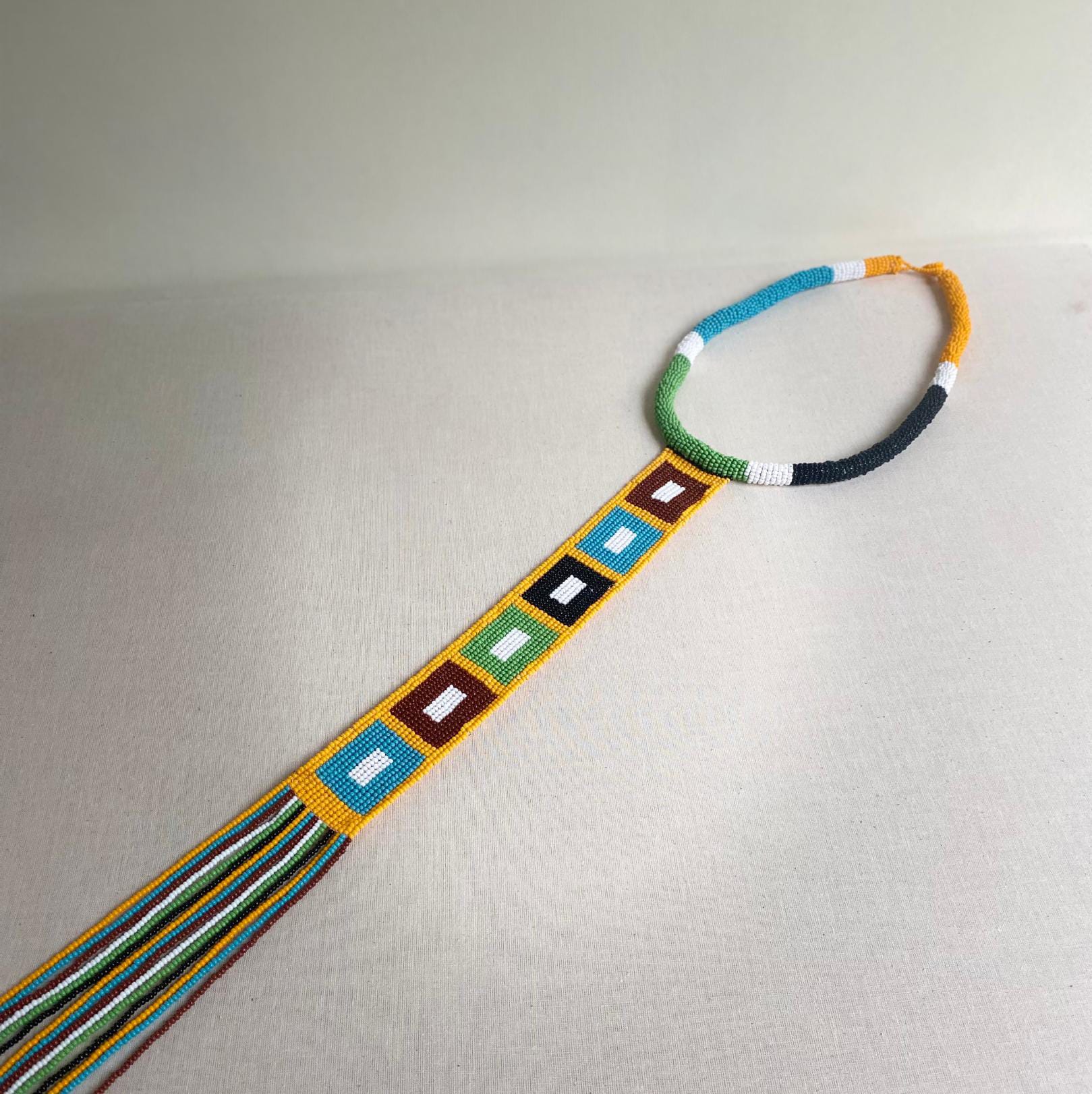 Beaded necklace with a long pendant, inspired by Contra.Joburg. The necklace features yellow, white, turquoise, orange, and black beads. The pendant shows coloured squares—blue, brown, green, yellow, red—echoing the Weight of Silence motif.