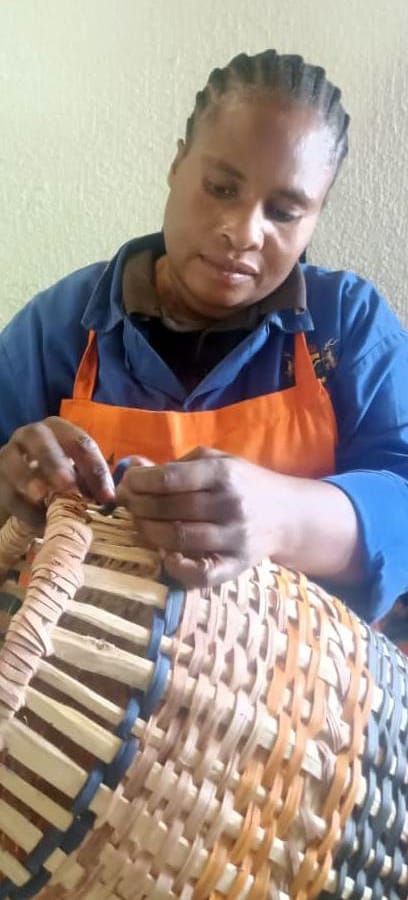A woman wearing a blue shirt and orange apron, part of Contra.Joburg, focuses on weaving a basket from light and dark natural materials, her hands moving with the weight of silence as she intricately intertwines the fibres in a softly lit setting.