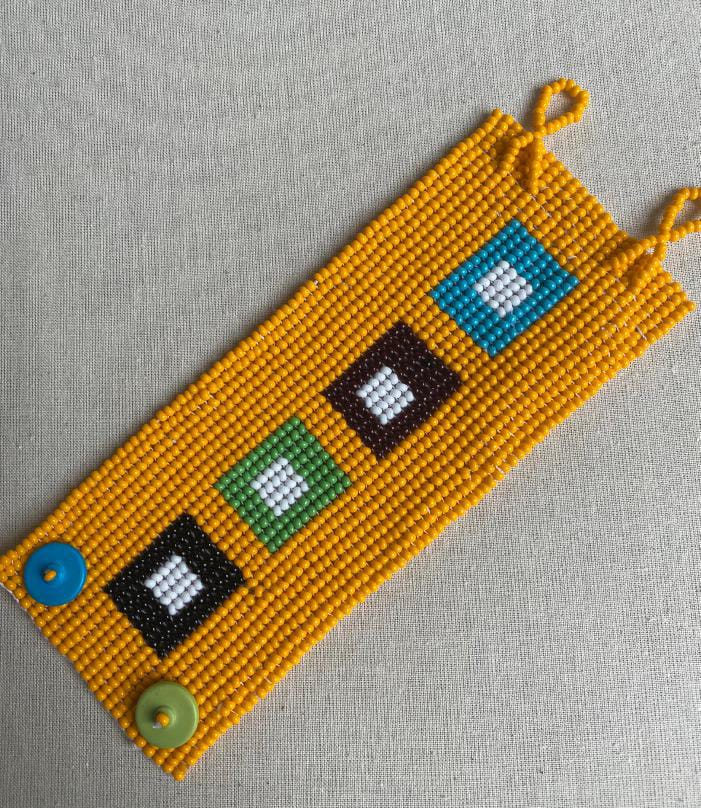 A handmade beaded bracelet with a yellow background and five square patterns in blue, black, brown, green, and black—each with a white centre. Inspired by Contra.Joburg’s Weight of Silence, it features blue and green buttons for fastening.
