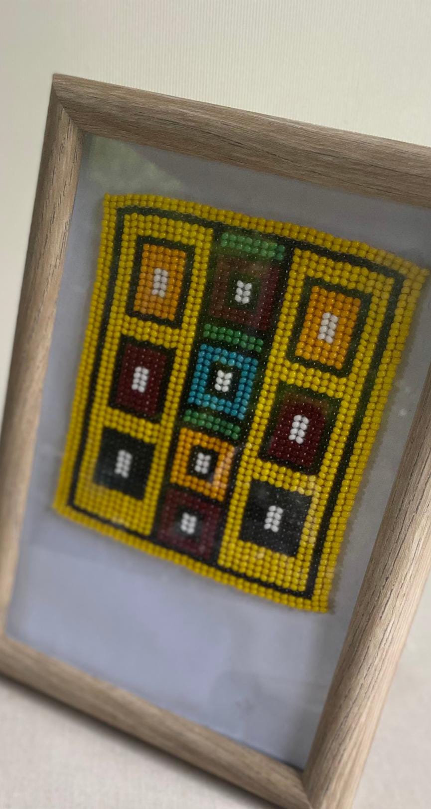 A framed beadwork art piece from Contra.Joburg features a yellow-bordered geometric pattern with colourful squares—red, green, brown, black, and white—displayed against a white background within a wooden frame, photographed at a slight angle.