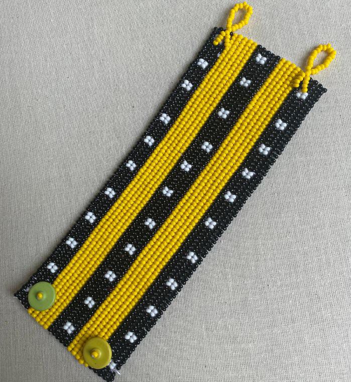 A beaded bracelet featuring alternating black and yellow vertical stripes, inspired by Contra.Joburg. The black stripes have small white bead clusters, whilst solid yellow beads and green buttons evoke the subtle Weight of Silence in its design.