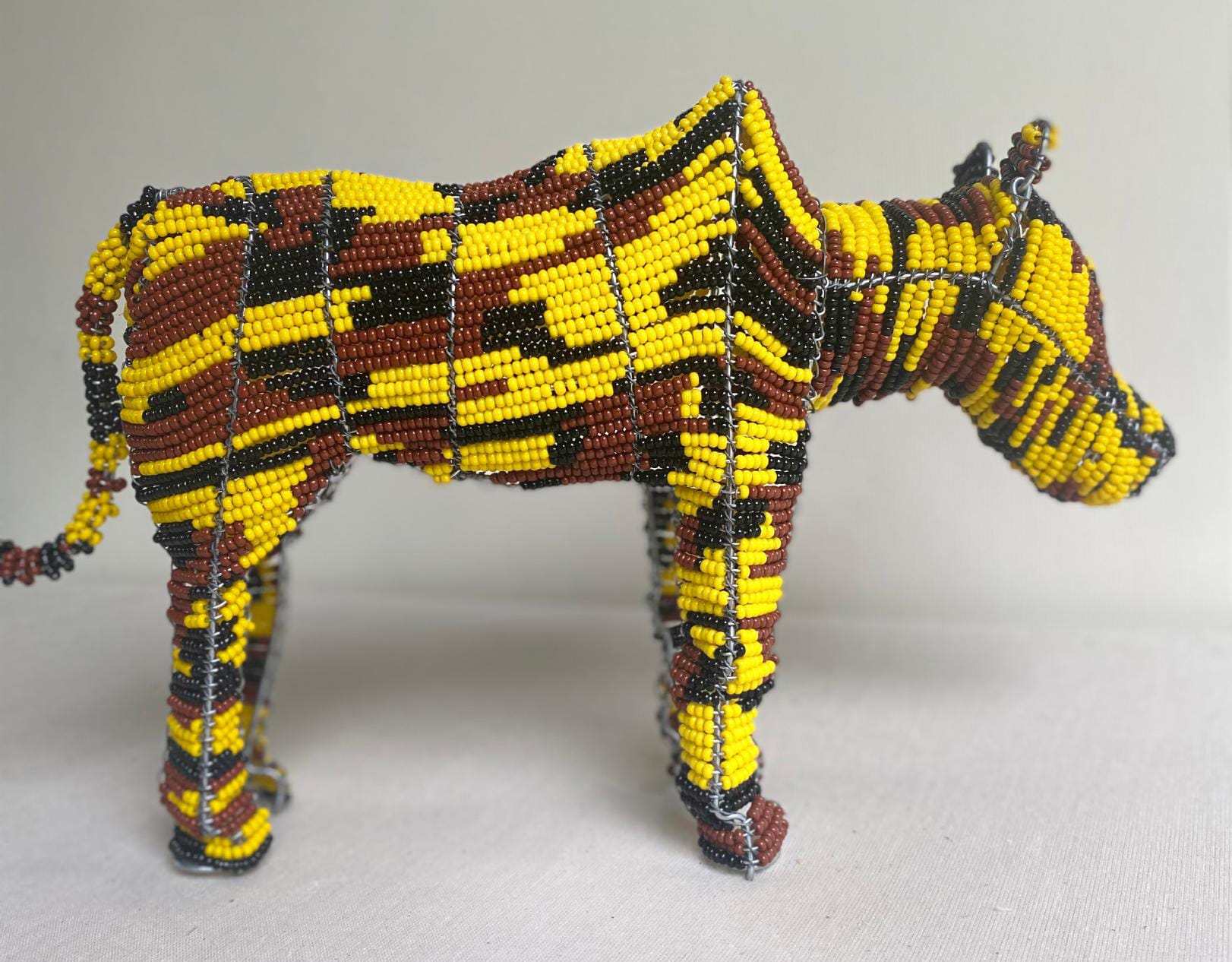 A beaded animal sculpture, possibly a zebra or horse, stands on a white surface. Part of the Contra.Joburg collection, its body is covered in small yellow, black, and brown beads in a patterned design. The background is plain and light-coloured.